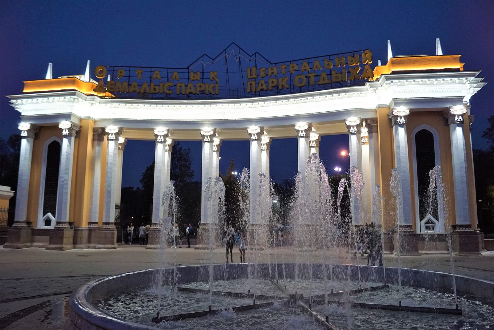 Подсвеченная колоннада главного входа Центрального парка Алматы с фонтаном ночью
